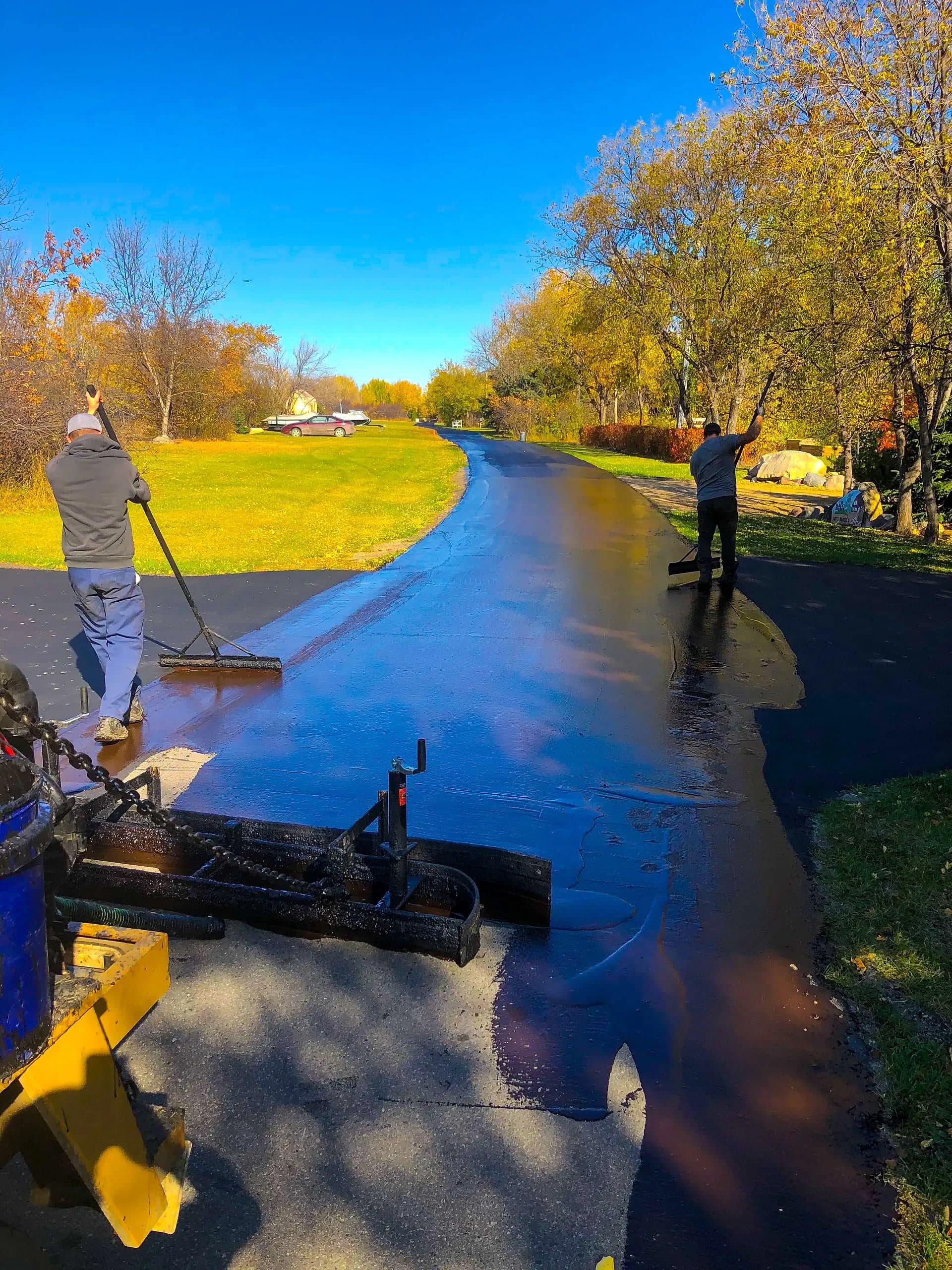 Crew spreading sealer across a long driveway with squeegees on a sunny fall day