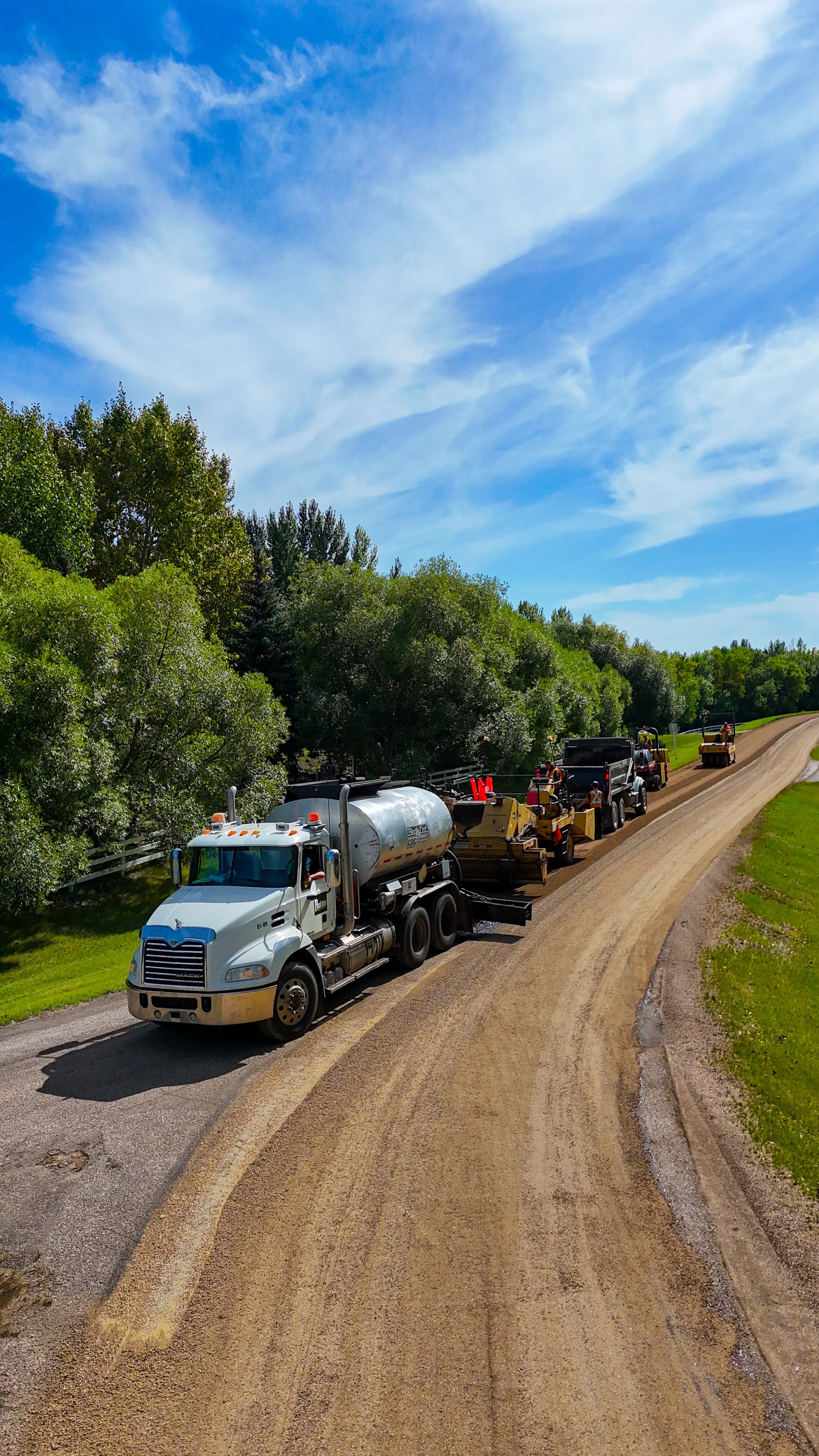 Raider Asphalt emulsion tanker leading the chip-seal train down a tree-lined road, one lane already sealed