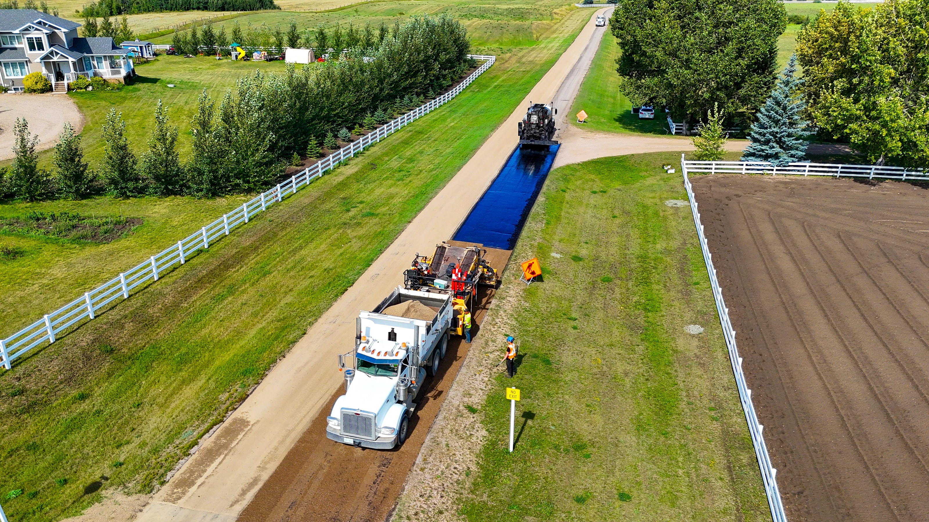 Aerial view of a Raider Asphalt crew laying fresh hot mix asphalt on a long acreage driveway lined with white fence