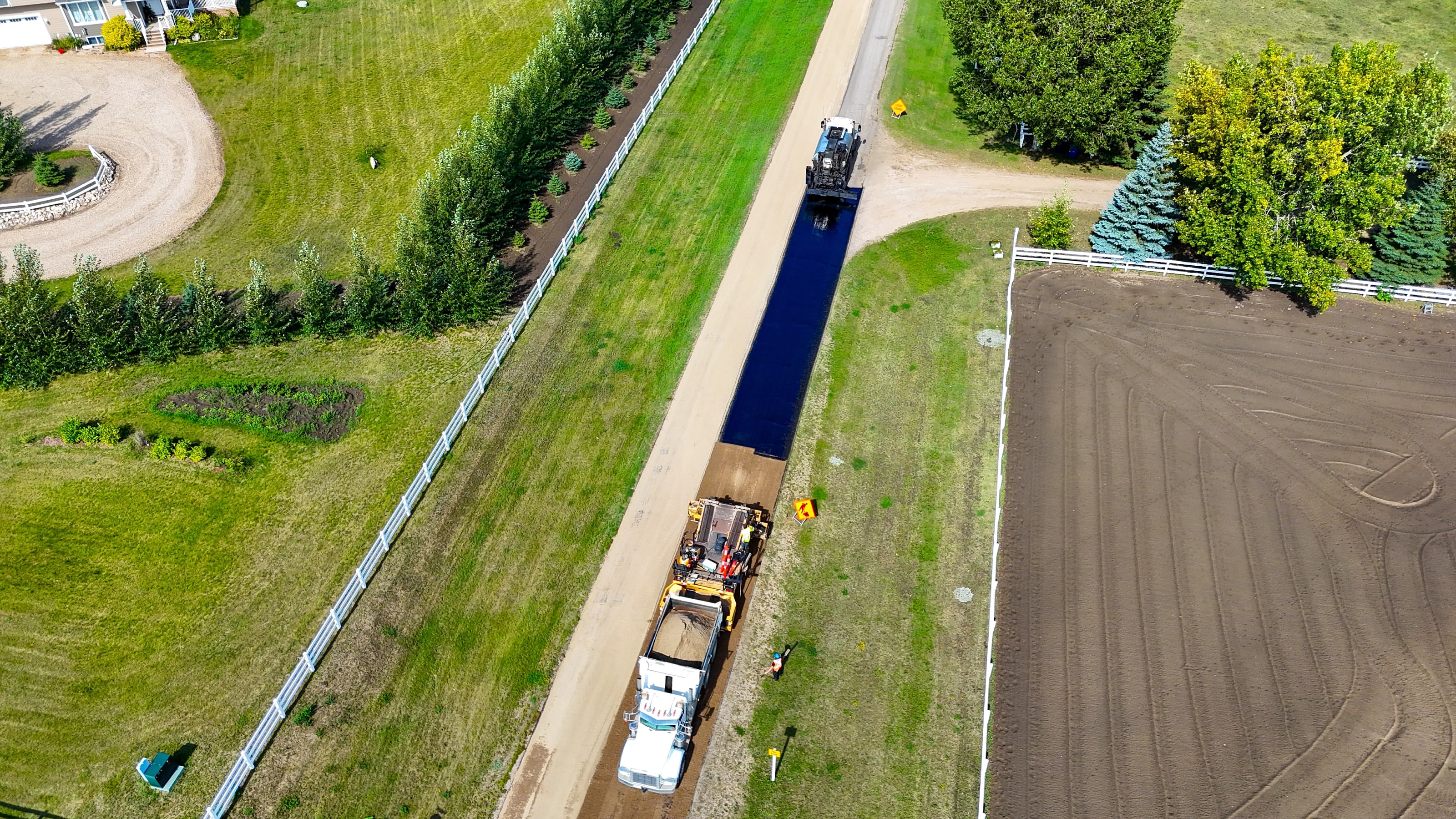 Drone view of a paver laying a strip of fresh black asphalt down an acreage driveway near Saskatoon