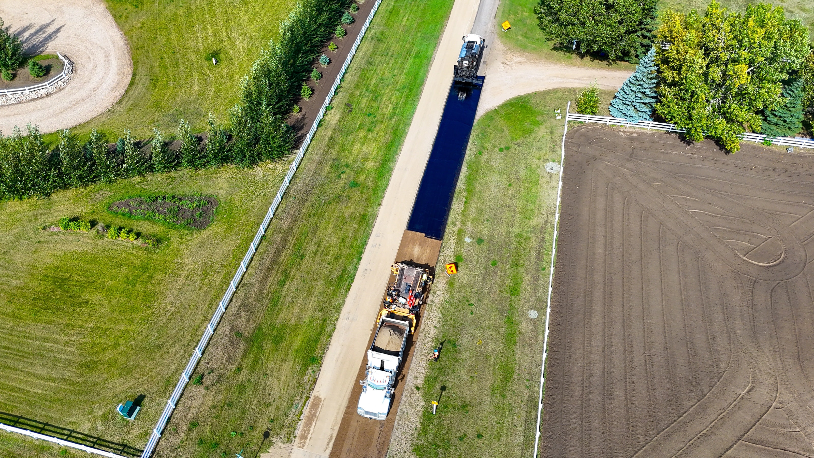 Fresh hot mix asphalt driveway taking shape between a white rail fence and a freshly tilled field, seen from above