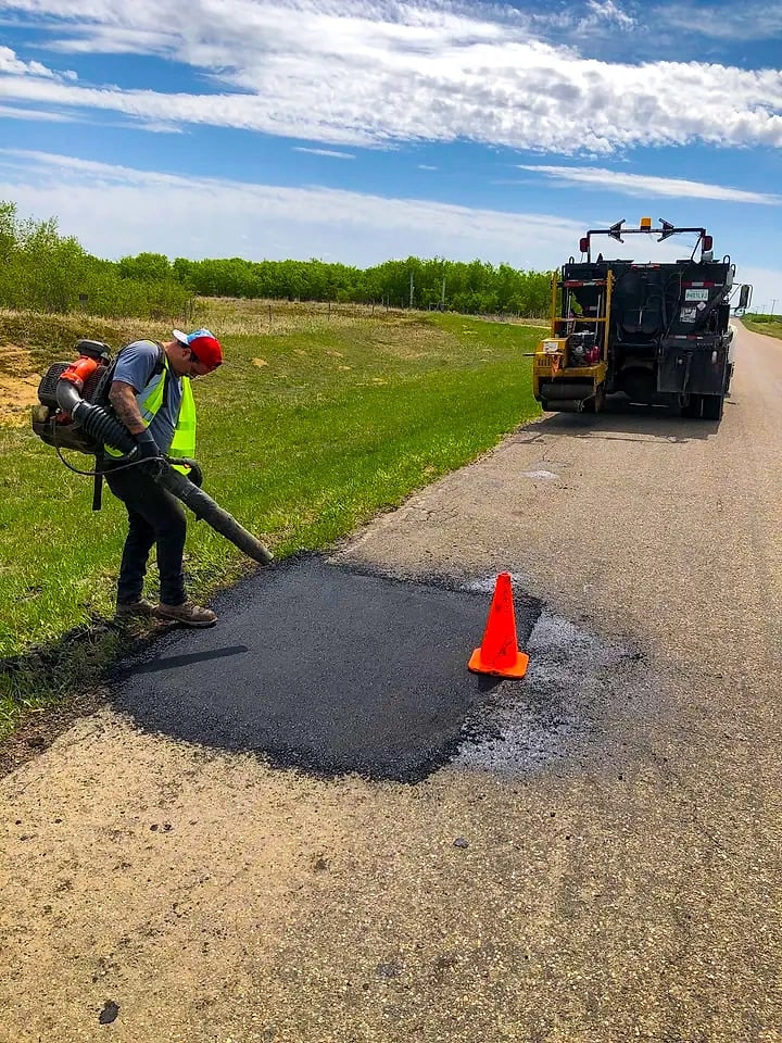 Crew member clearing debris from a fresh hot mix asphalt patch on a rural road